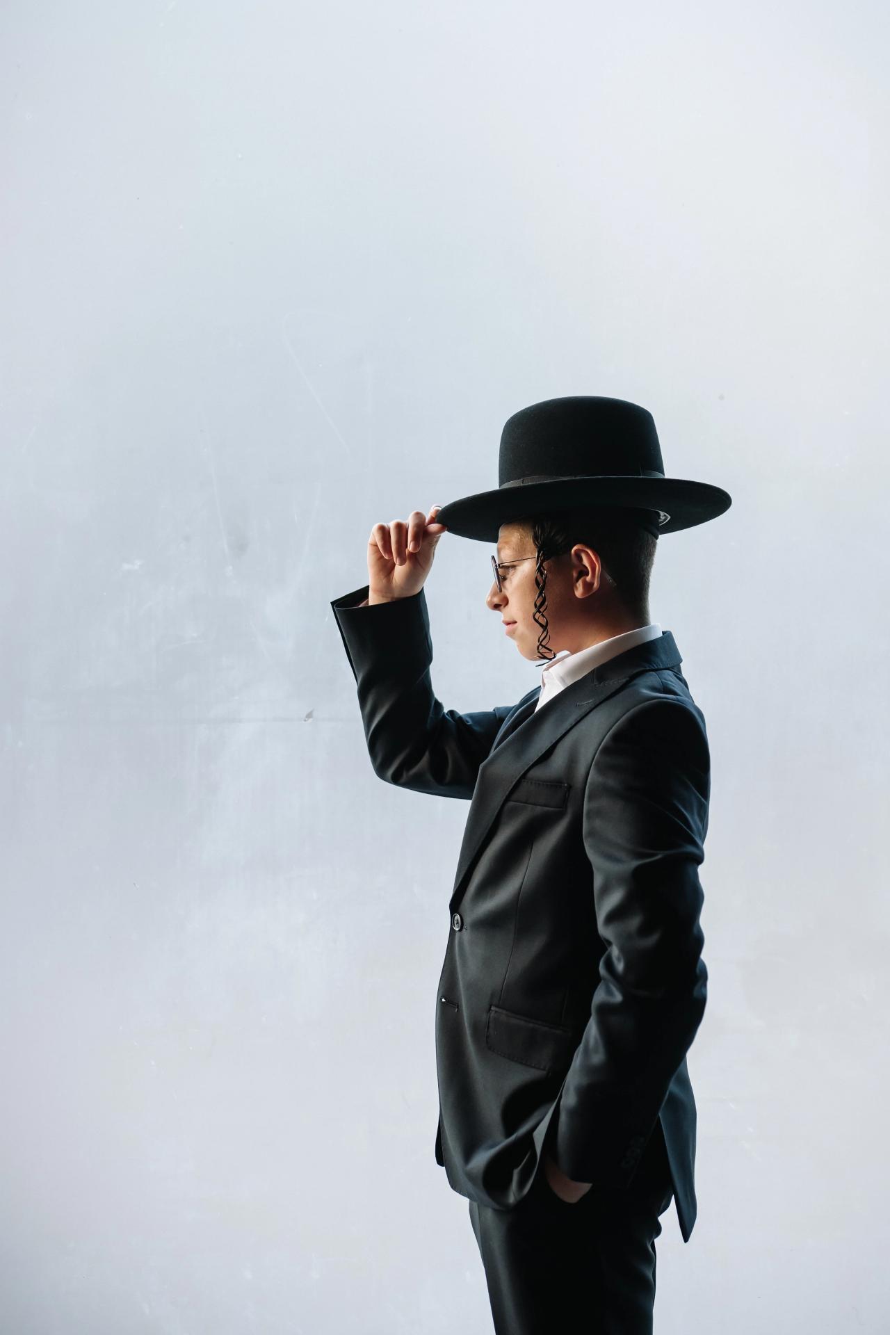 Bar Mitzvah boy in a dark suit and black hat tipping his hat, standing against a bright plain wall. Bar Mitzvah photography at The Arches, Brooklyn, NY.