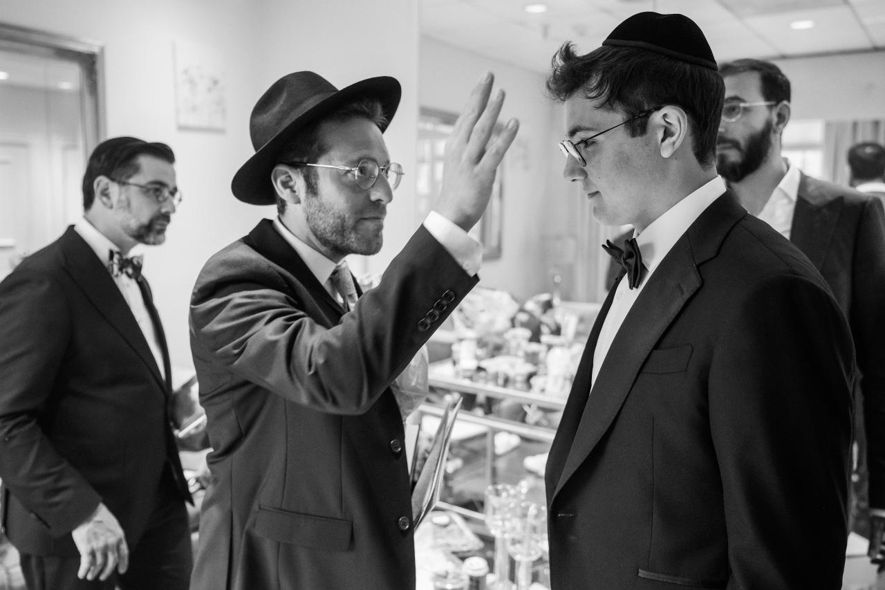 Black-and-white candid moment of a suited guest gesturing and the groom smiling as they stand face-to-face at an indoor celebration, with other guests in the background. Wedding photography at Falkirk Estate Country Club, Central Valley, NY.