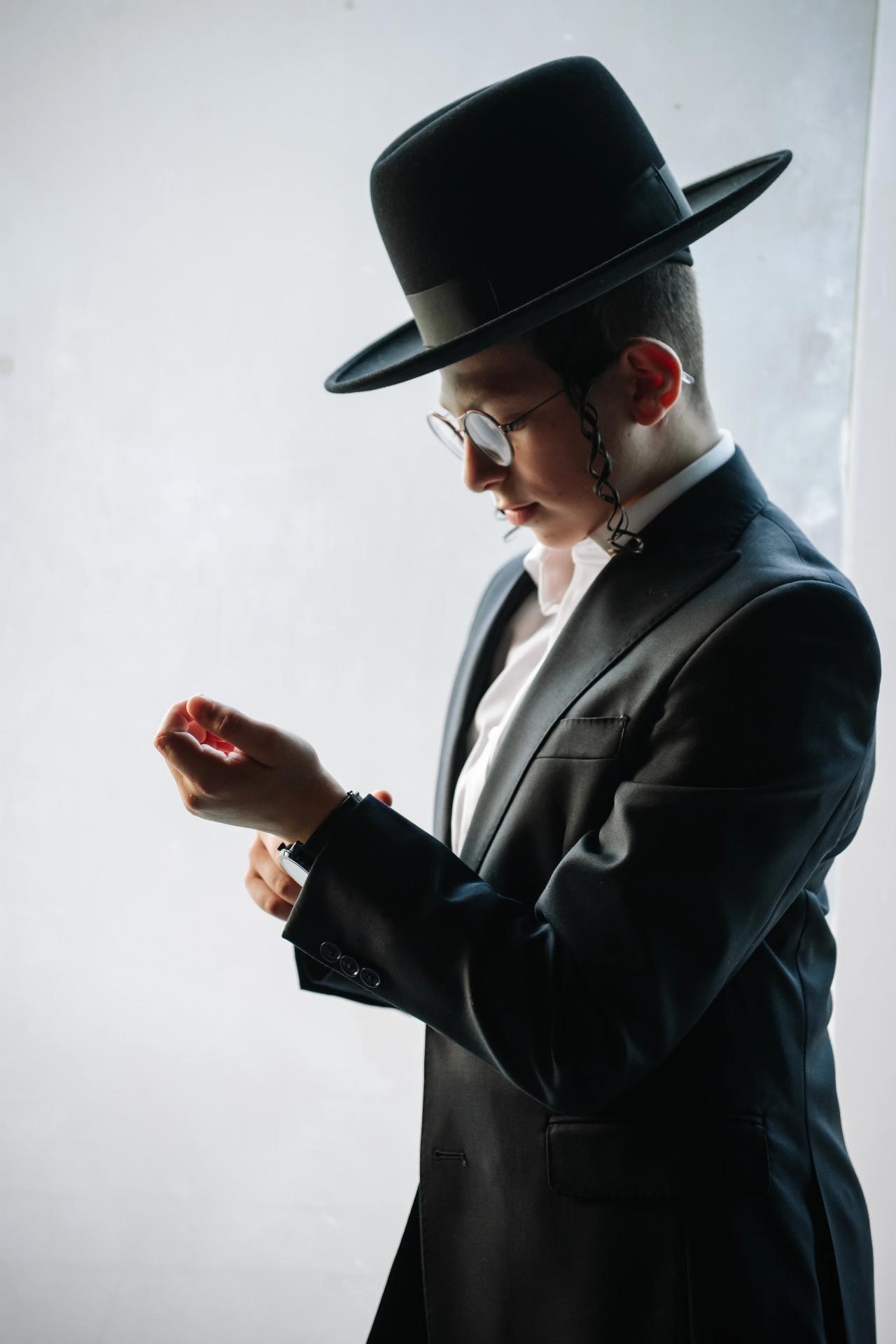 Close side-profile portrait of the Bar Mitzvah boy in a black hat and suit, looking down at his hands against a bright background. Bar Mitzvah photography at The Arches, Brooklyn, NY.