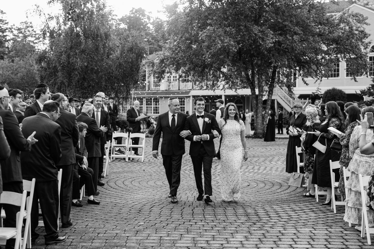 Black-and-white photo of a couple walking down an outdoor wedding aisle as confetti falls, with guests seated along both sides. Wedding photography at Falkirk Estate Country Club, Central Valley, NY.