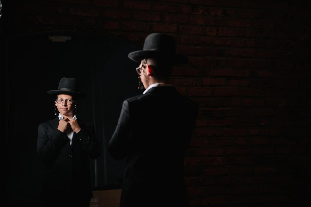 Bar Mitzvah boy in a black hat and suit adjusting his collar in a mirror beside a brick wall in dramatic low light. Bar Mitzvah photography at The Arches, Brooklyn, NY.