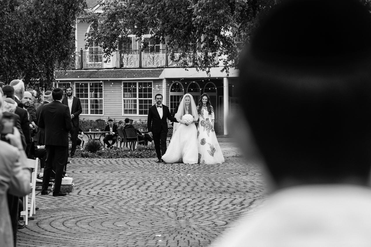 Black-and-white outdoor ceremony photograph of a bride and groom walking down a cobblestone aisle toward the chuppah area, framed by trees and building windows. Wedding photography at Falkirk Estate Country Club, Central Valley, NY.