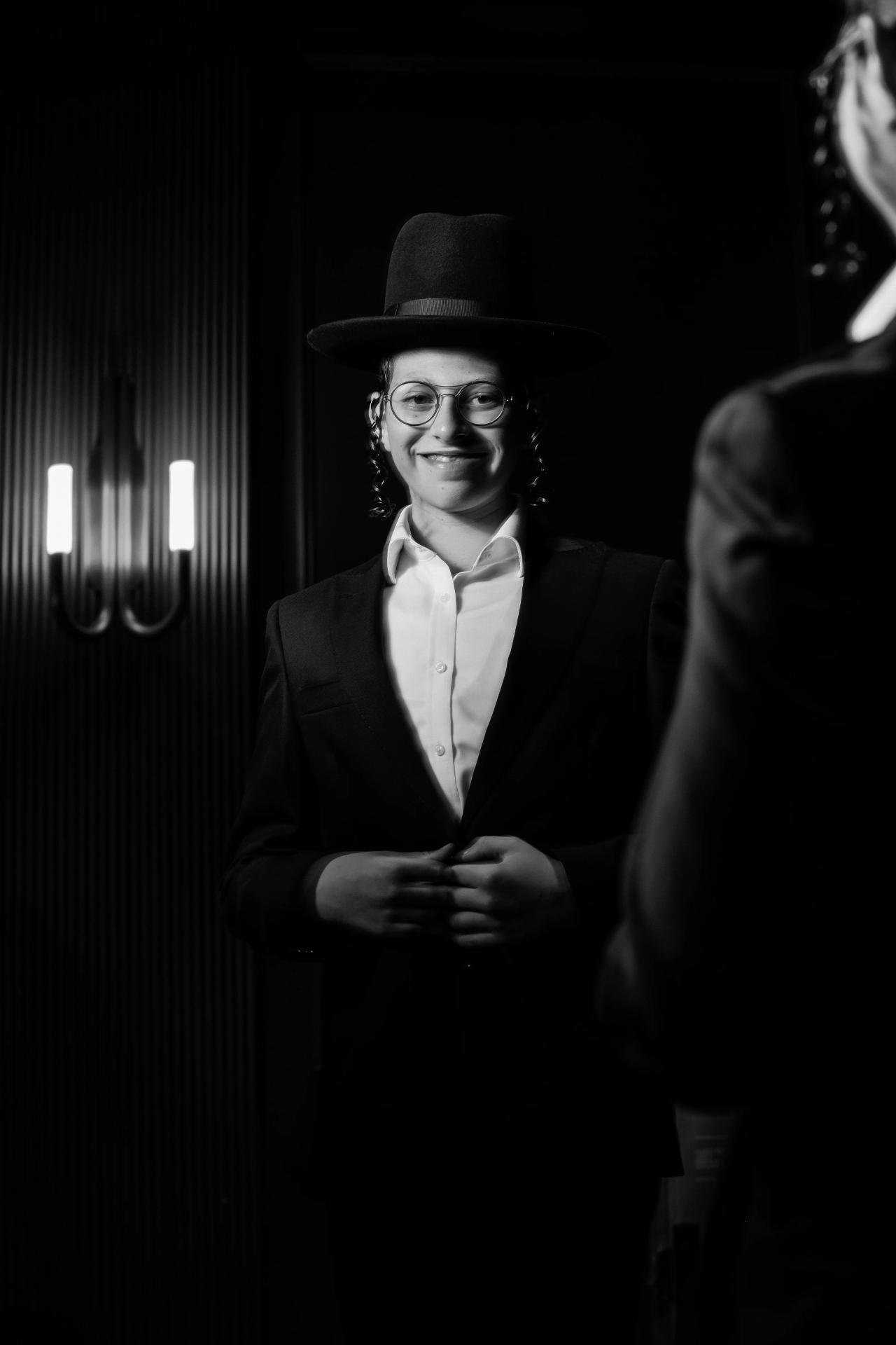 Black-and-white portrait of a smiling guest wearing a dark hat and formal jacket, standing against a ribbed wall with soft wall lighting. Bar Mitzvah photography at The Arches, Brooklyn, NY.