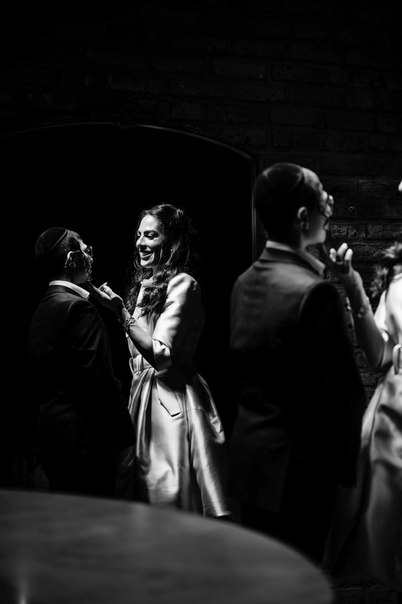 Black-and-white candid of a smiling woman in a satin dress laughing during the party, facing a guest in a dark suit near a table; brick wall backdrop with guests blurred behind them. Bar Mitzvah photography at The Arches, Brooklyn, NY.