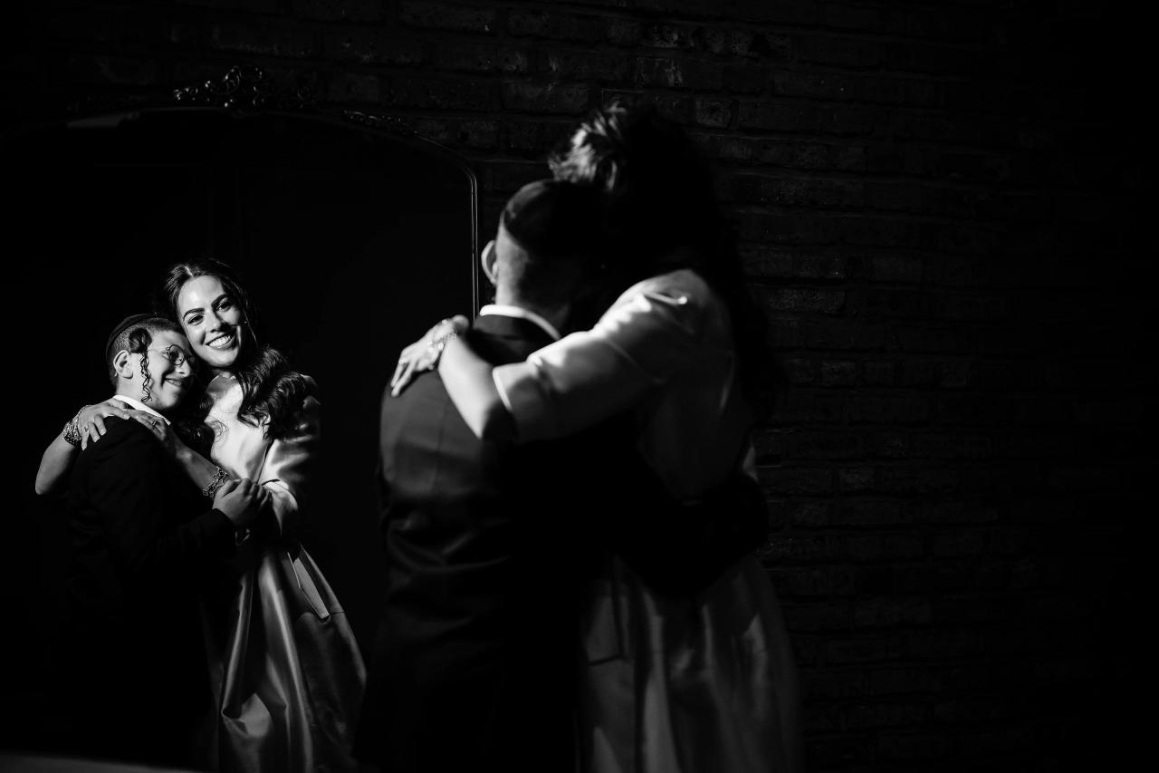 Black-and-white candid of a joyful dance-floor hug: a smiling woman and child embrace each other while a man approaches from the foreground in a dark brick room. Bar Mitzvah photography at The Arches, Brooklyn, NY.