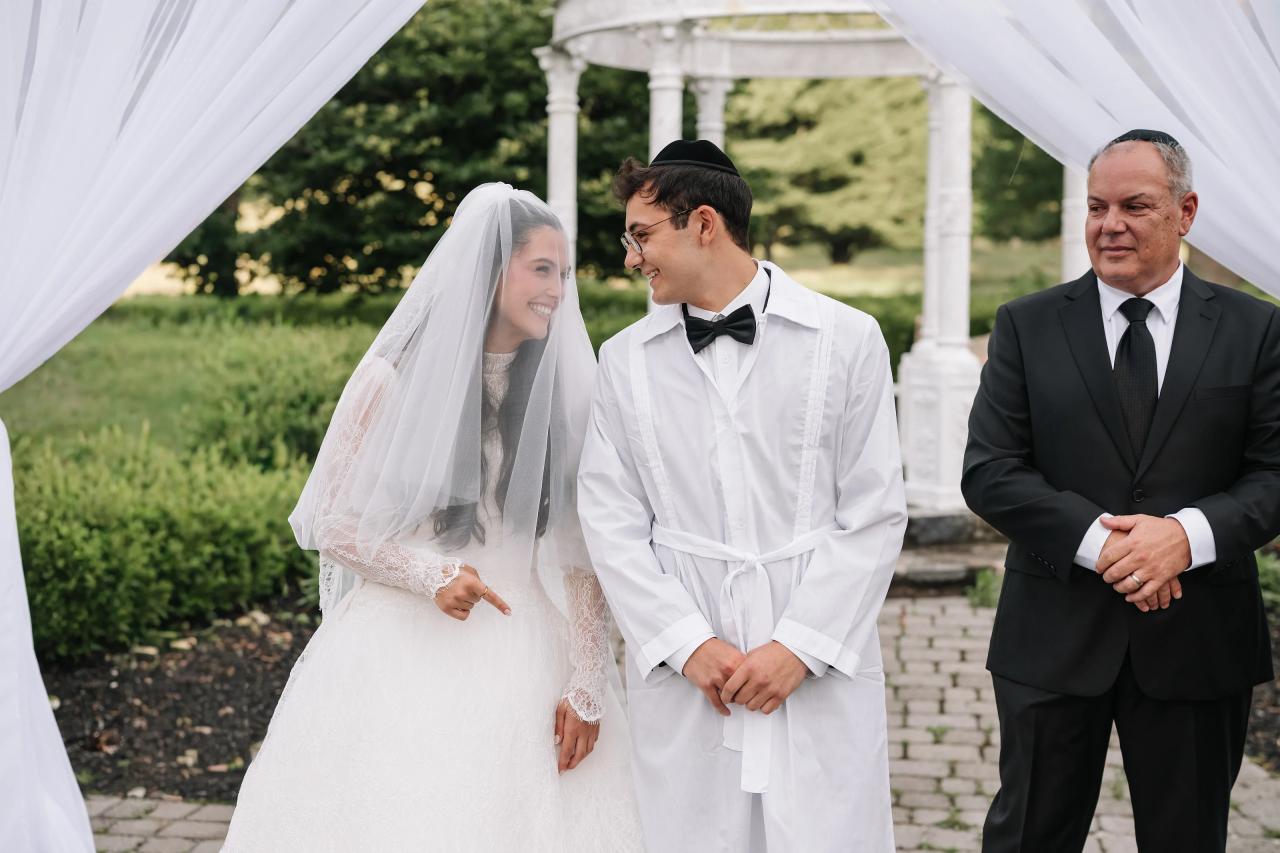 Bride and groom face each other under a white draped canopy during badeken, with the groom in traditional white attire and a man in a dark suit standing nearby. Wedding photography at Falkirk Estate Country Club, Central Valley, NY.