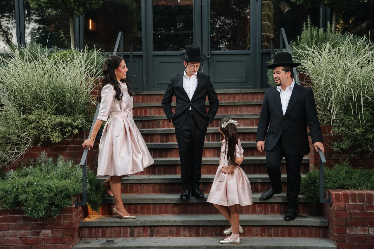 Bar Mitzvah boy and guests in formalwear on brick steps outdoors, surrounded by greenery. Bar Mitzvah photography at The Arches, Brooklyn, NY.