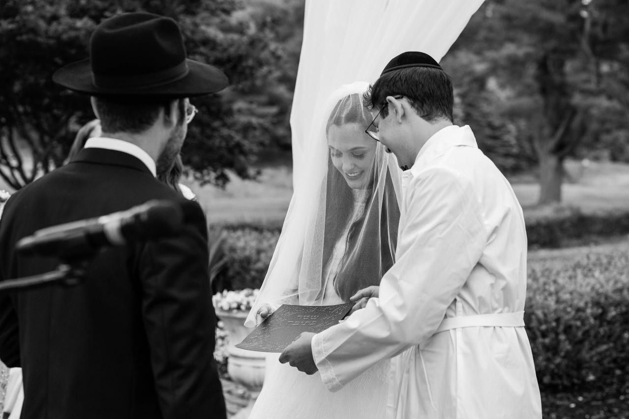 Black-and-white photo of a groom leaning close to his bride during badeken as she smiles under a veil, hands gently extended outdoors with hedges and greenery in the background. Wedding photography at Falkirk Estate Country Club, Central Valley, NY.