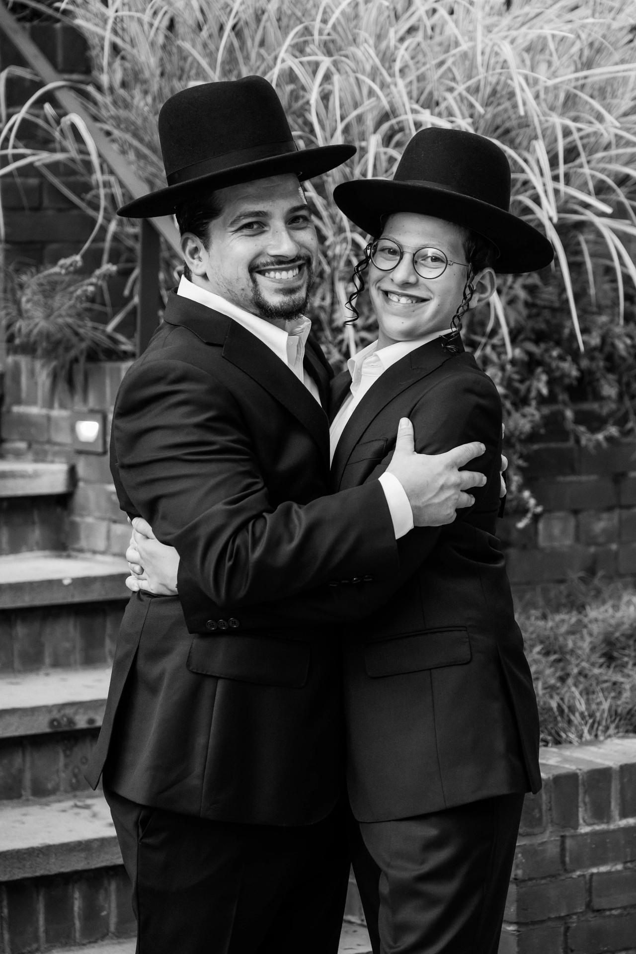 Black-and-white portrait of the Bar Mitzvah boy smiling in an embrace with another guest, with grasses and a brick wall behind them. Bar Mitzvah photography at The Arches, Brooklyn, NY.