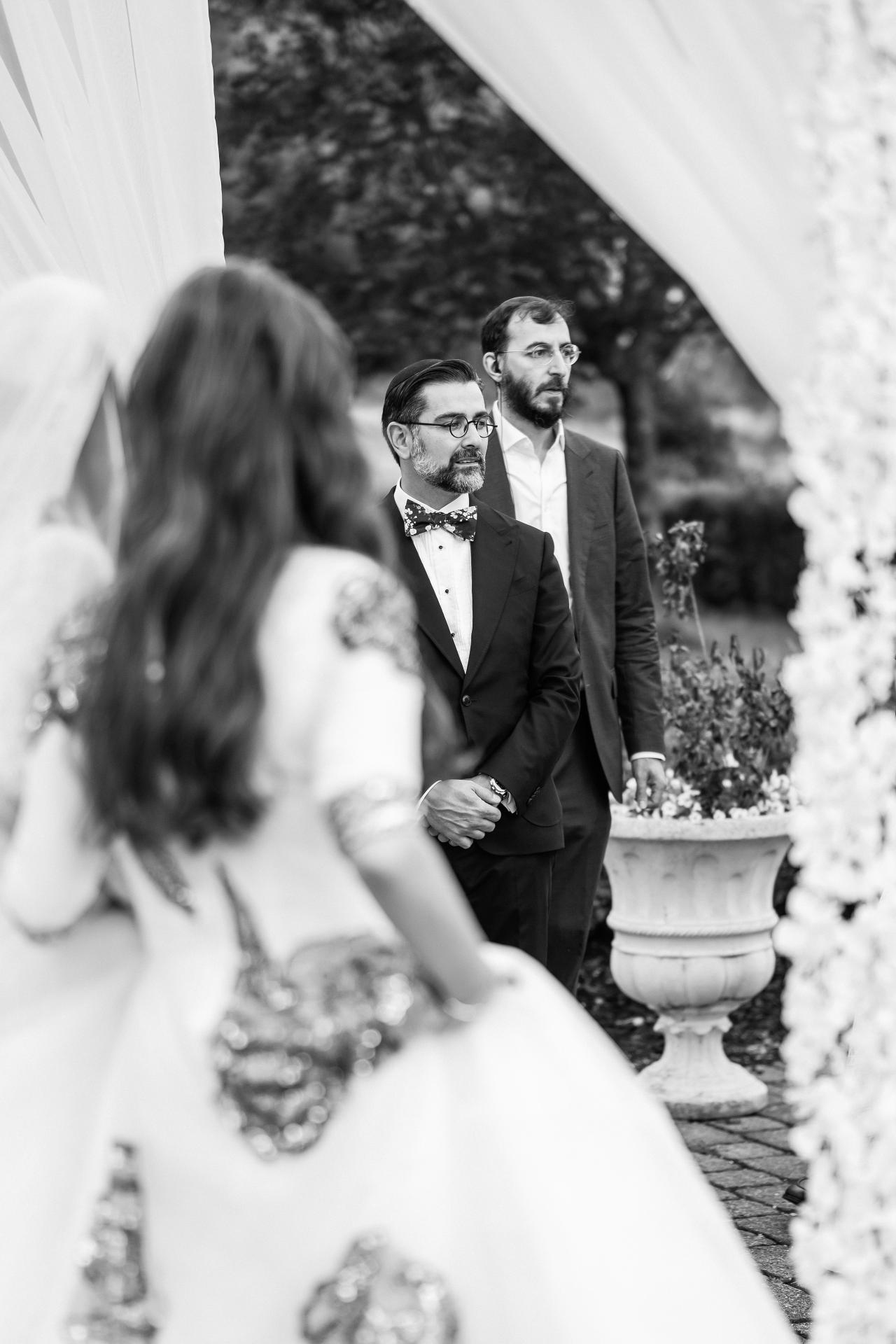 Black-and-white ceremony photo with a groom in a suit standing at mid-ground while the bride is out of focus in the foreground; flowers and chuppah structure frame the scene. Wedding photography at Falkirk Estate Country Club, Central Valley, NY.