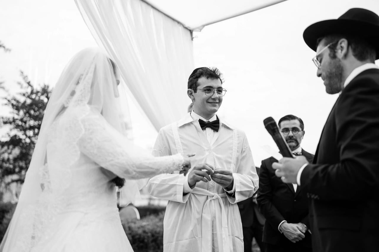 Black-and-white wedding ceremony photo under a white canopy: the groom stands smiling as the bride’s veil reaches across the frame; a rabbi and guests are partially visible at the sides. Wedding photography at Falkirk Estate Country Club, Central Valley, NY.