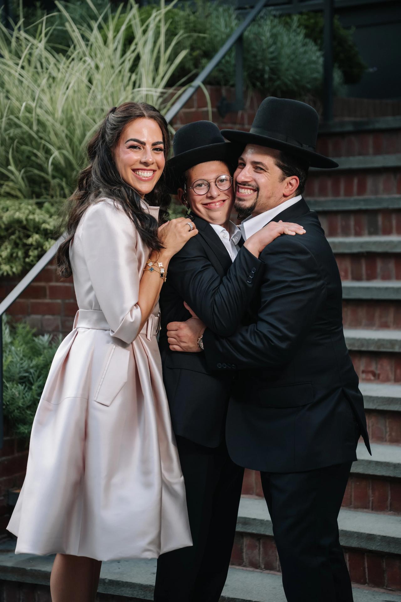 Bar Mitzvah boy smiling in an embrace with two adults outdoors near brick and greenery. Bar Mitzvah photography at The Arches, Brooklyn, NY.