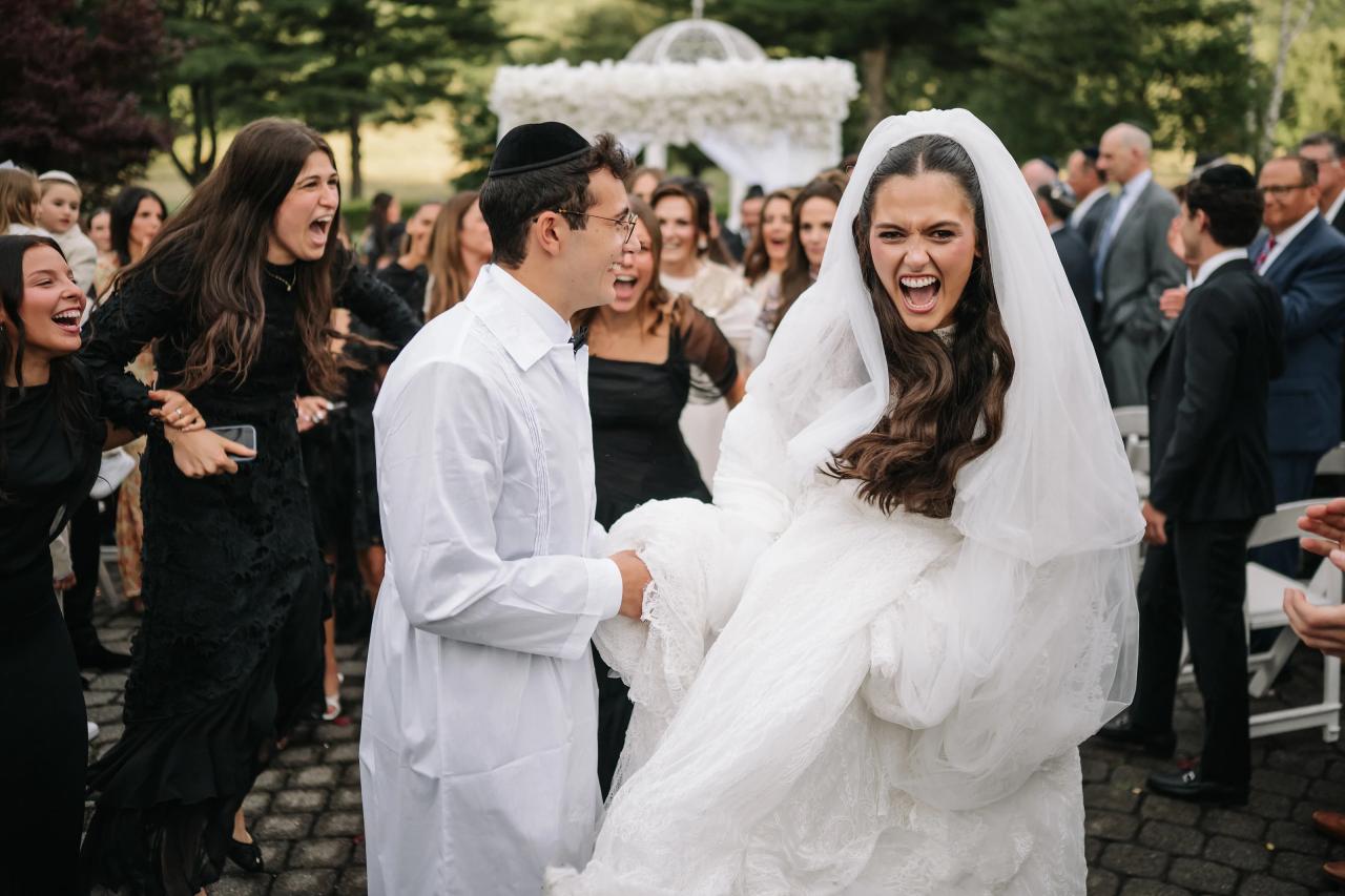 Bride and groom laughing and embracing outdoors with the crowd cheering during the celebration, white veil and suit against a green garden background. Wedding photography at Falkirk Estate Country Club, Central Valley, NY.
