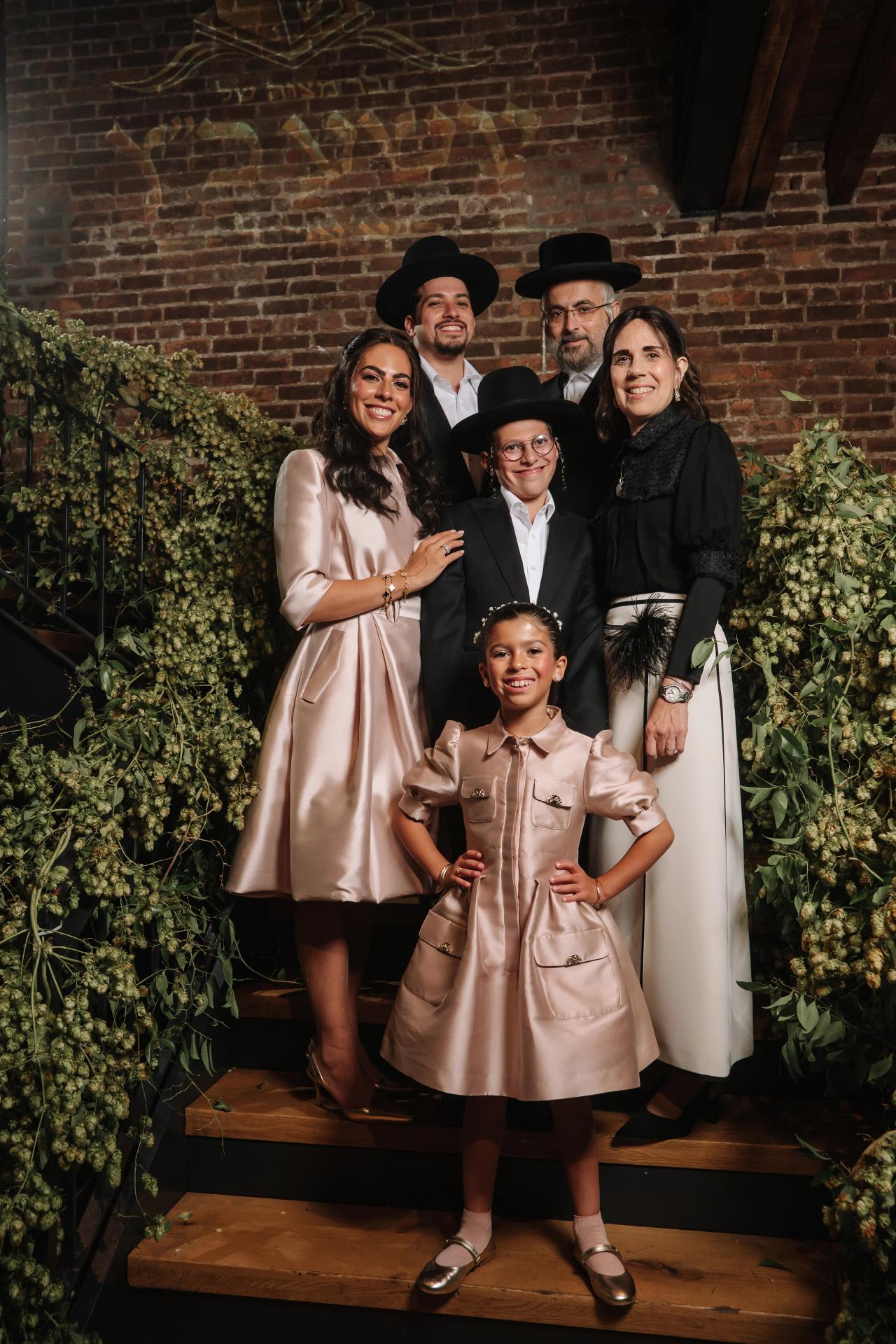 Smiling group of family members dressed in formal black hats and satin outfits, posed on tiered steps in front of lush green floral arrangements against a brick wall. Bar Mitzvah photography at The Arches, Brooklyn, NY.