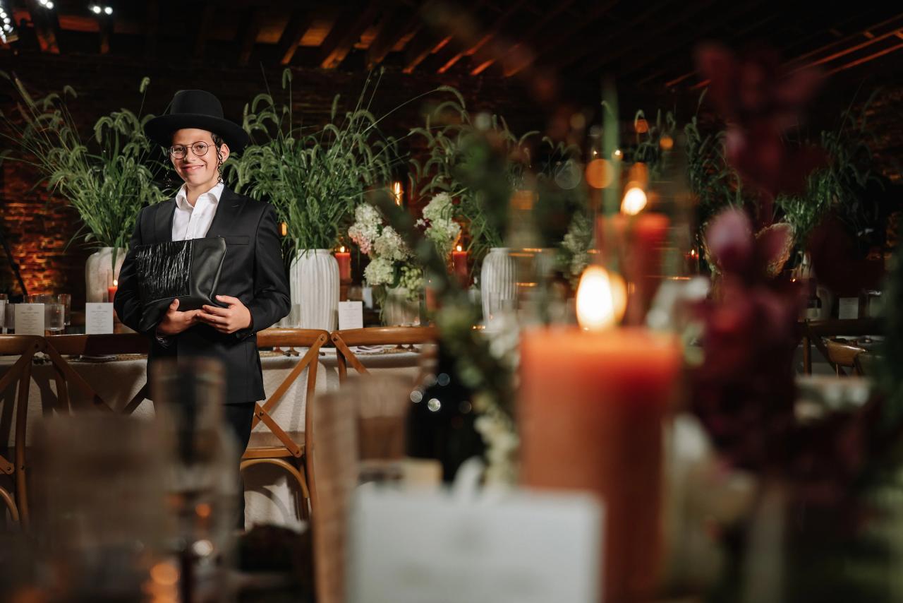 Young guest in black formal attire wearing a hat and glasses standing in front of floral centerpieces and candlelit reception tables, with soft bokeh lights in the background. Bar Mitzvah photography at The Arches, Brooklyn, NY.