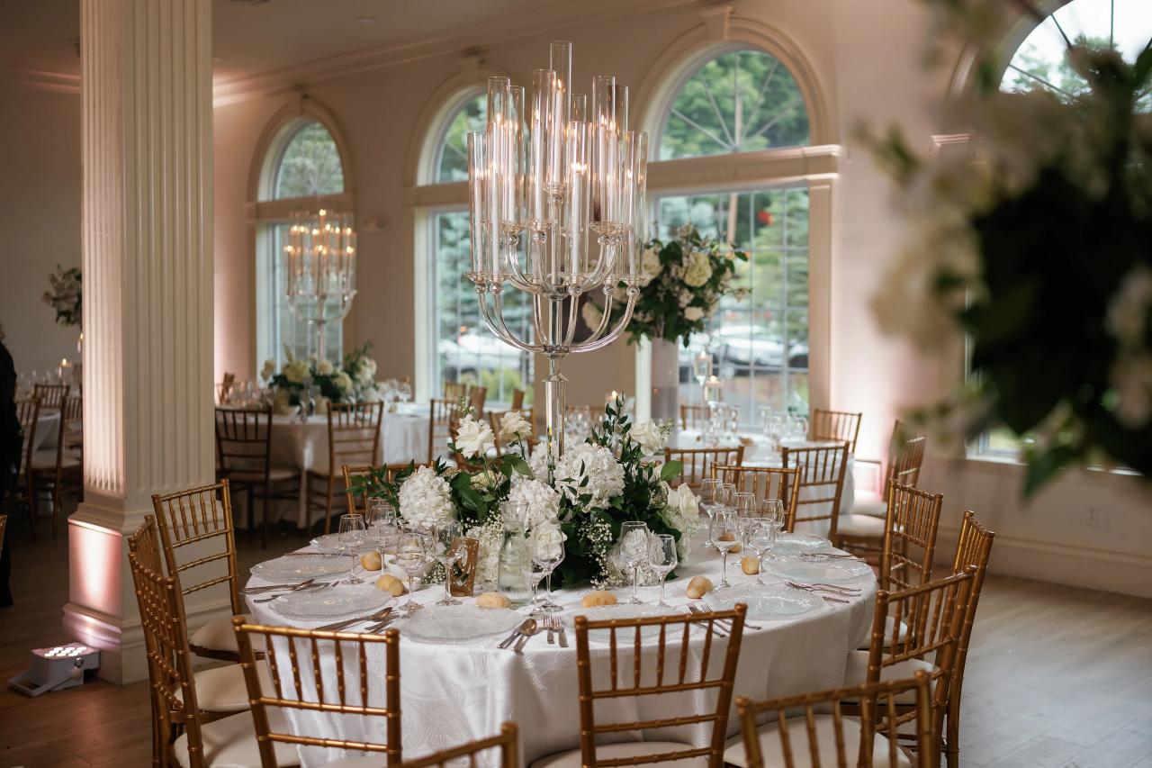 Styled reception table with floral centerpiece and tall candelabra under a chandelier in a bright arched event hall. Wedding photography at Falkirk Estate Country Club, Central Valley, NY.