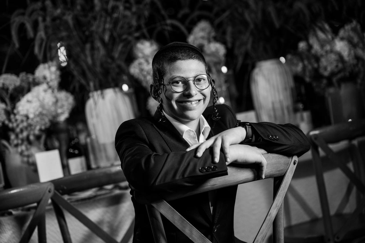 Black-and-white portrait of a smiling young boy seated at a banquet chair with floral centerpieces and guests softly blurred in the background. Bar Mitzvah photography at The Arches, Brooklyn, NY.