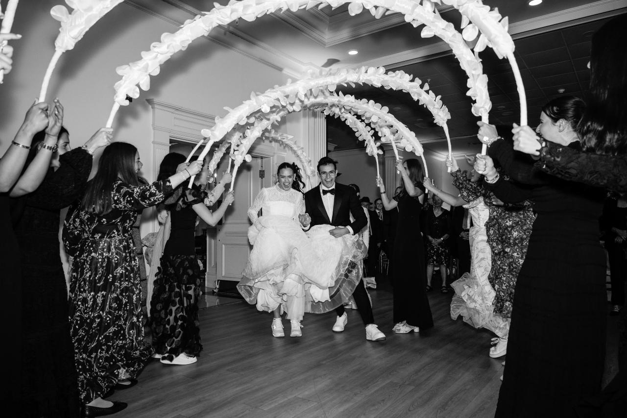Black-and-white wedding dance with guests holding a floral canopy overhead while the bride and groom walk forward through confetti, surrounded by joyful raised hands. Wedding photography at Falkirk Estate Country Club, Central Valley, NY.