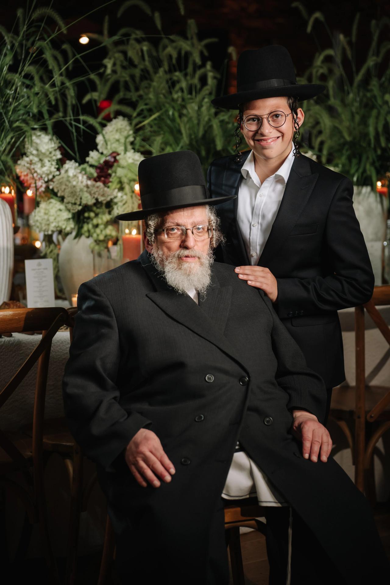 Two seated men in black hats at a candlelit event, one smiling with an arm around the other’s shoulder; floral décor and warm lights in the background. Bar Mitzvah photography at The Arches, Brooklyn, NY.