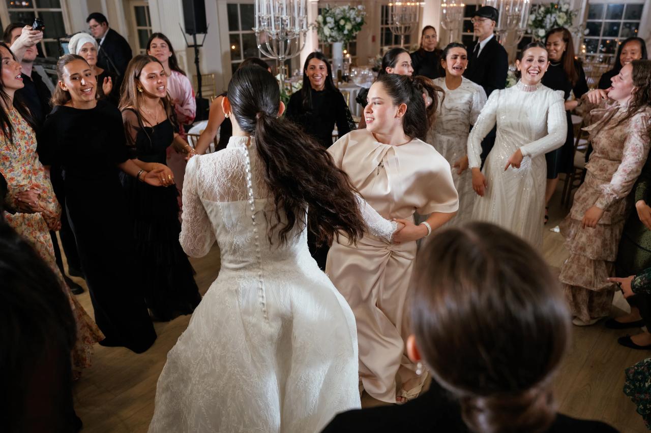 Candid dance-floor moment at a Jewish wedding: two guests laughing and moving while people around them clap and cheer, with the bride in the foreground turning toward the action. Wedding photography at Falkirk Estate Country Club, Central Valley, NY.