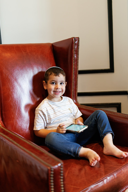 Smiling toddler seated on a red leather chair, holding a small booklet and looking toward the camera indoors.