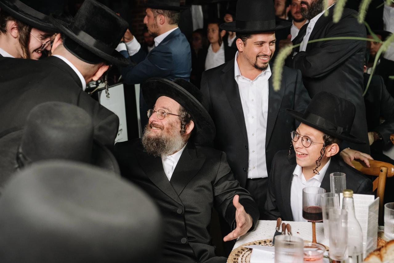 A warmly lit Bar Mitzvah reception scene with a bearded man in a black hat smiling and gesturing, seated at a table beside a young man wearing glasses and a hat, surrounded by guests in formal attire. Bar Mitzvah photography at The Arches, Brooklyn, NY.