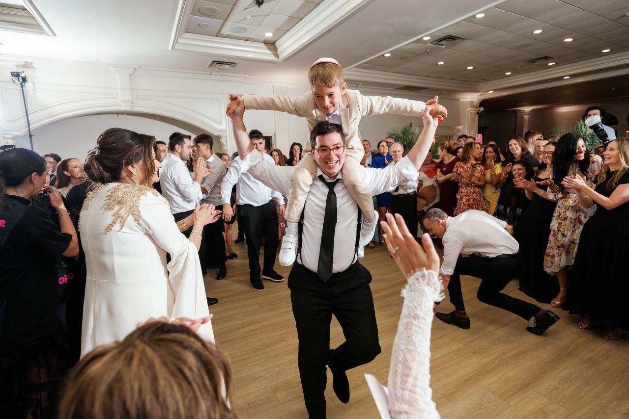 Wedding reception dance floor moment: the groom lifts a child’s arms while guests clap and smile in a bright ballroom. Wedding photography at Falkirk Estate Country Club, Central Valley, NY.