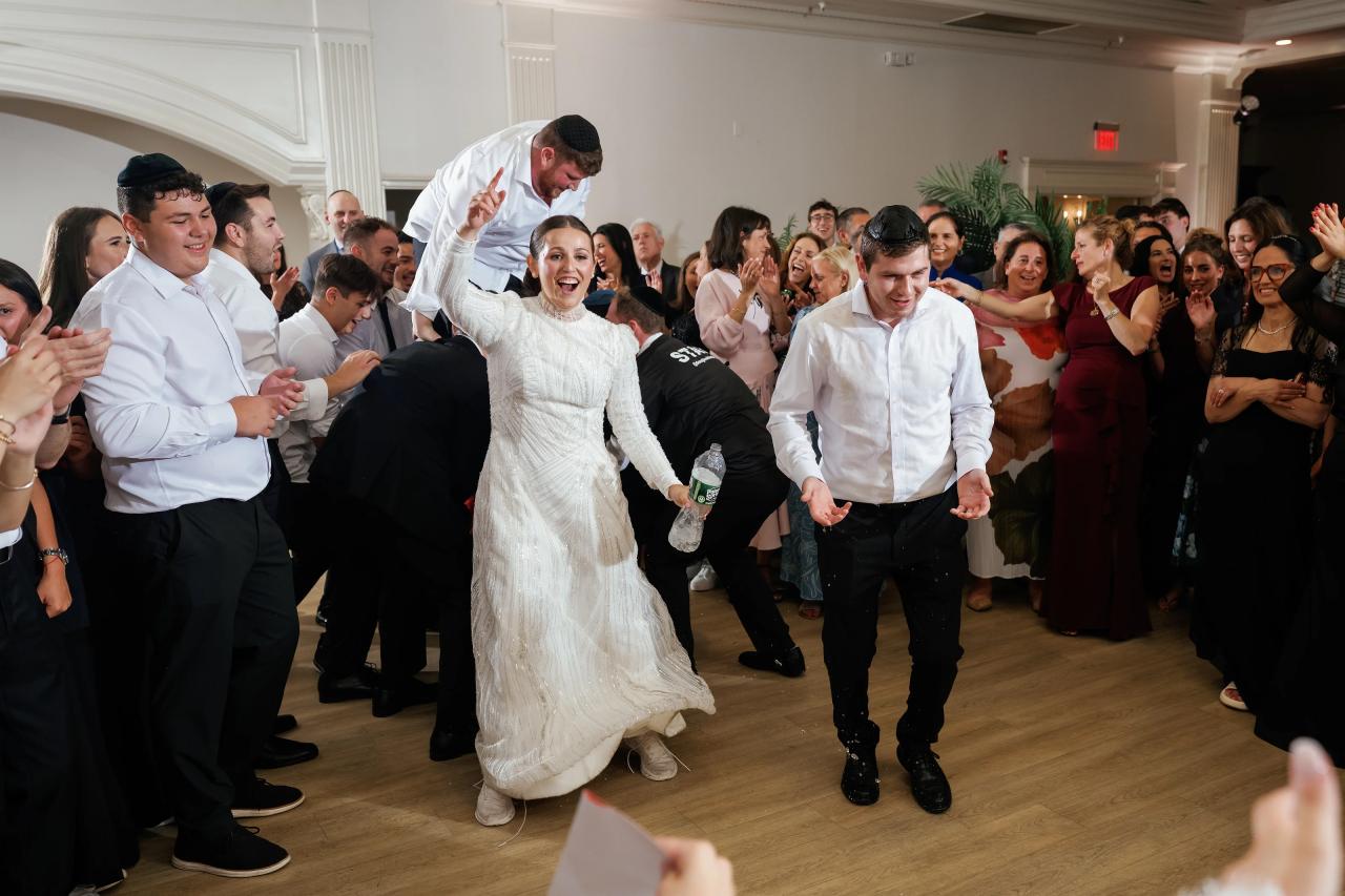 Bride and groom dancing at a wedding reception, surrounded by guests clapping and cheering in a brightly lit hall with white columns. Wedding photography at Falkirk Estate Country Club, Central Valley, NY.