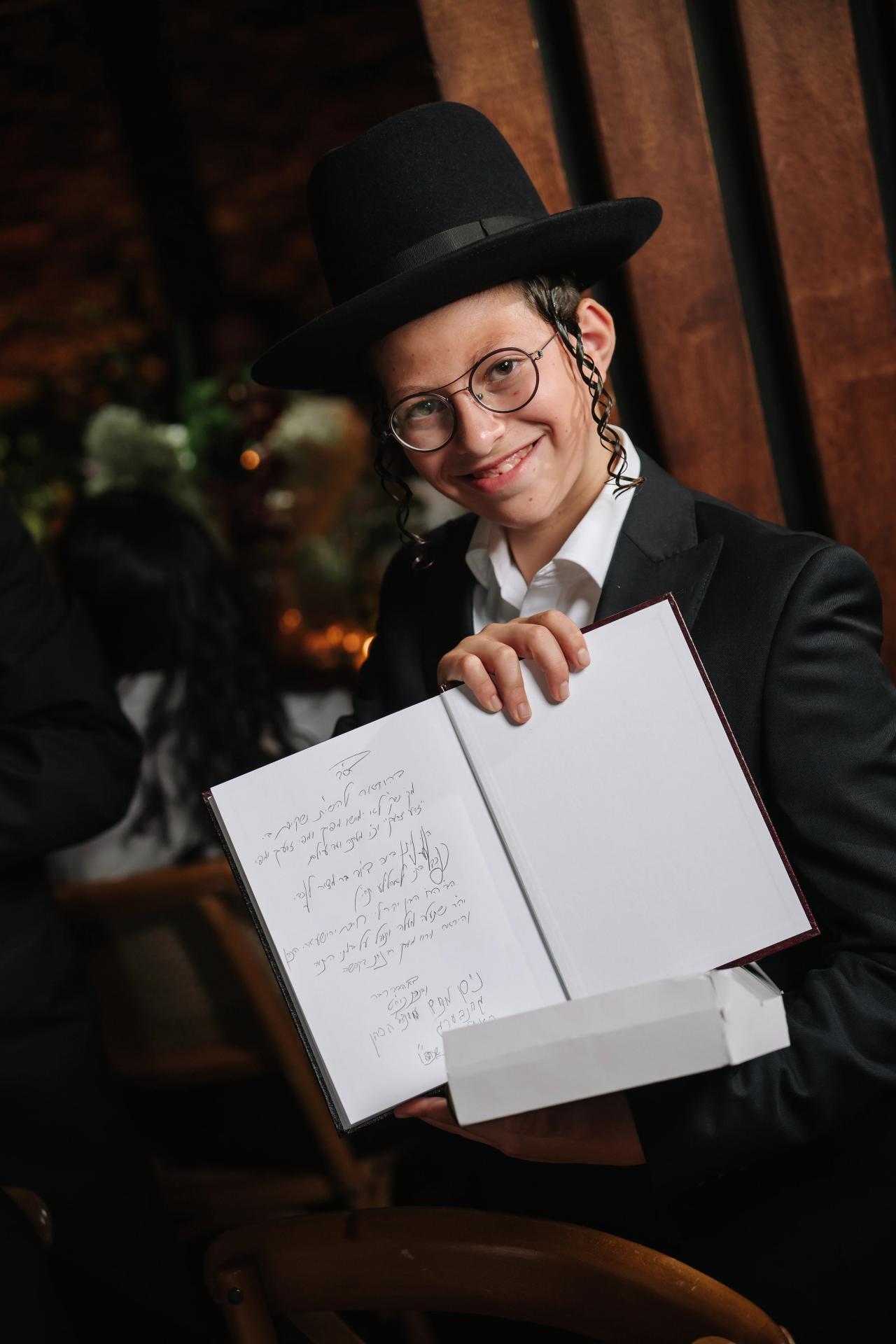 Smiling mitzvah guest in a black hat and suit holding an open certificate booklet, seated indoors with warm wood-toned background blur. Bar Mitzvah photography at The Arches, Brooklyn, NY.