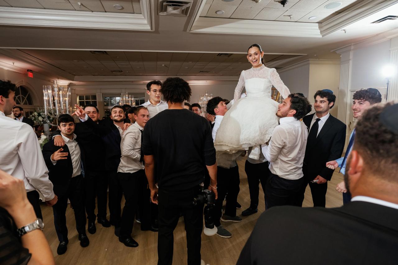 Bride in a white gown standing elevated above a cheering crowd during the reception, surrounded by guests in suits in a brightly lit indoor hall. Wedding photography at Falkirk Estate Country Club, Central Valley, NY.