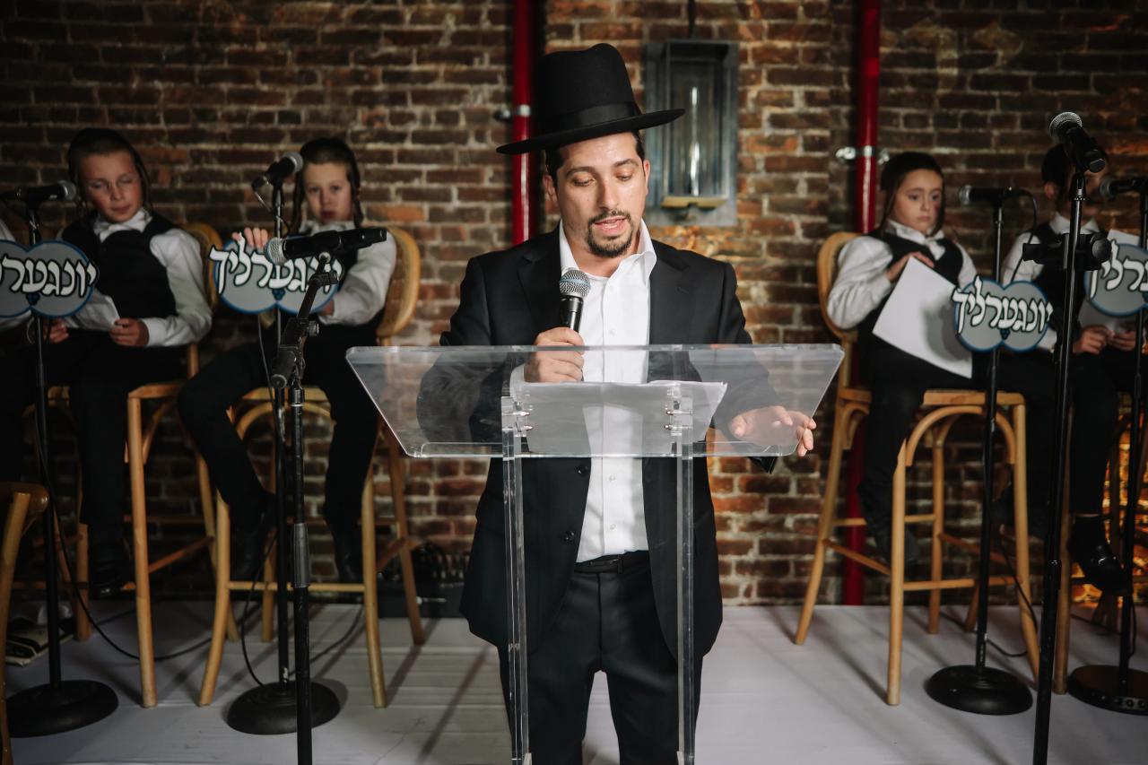 A man in a black hat and suit speaks at a clear podium with microphones while children in coordinated outfits sit behind him holding Hebrew signs. Bar Mitzvah photography at The Arches, Brooklyn, NY.