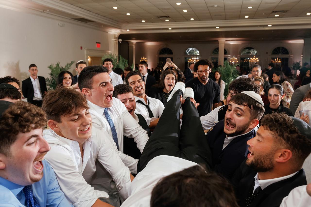 A lively crowd of guests in formal attire laughing and reacting together during a wedding celebration, captured from inside the seated dance-party moment. Wedding photography at Falkirk Estate Country Club, Central Valley, NY.