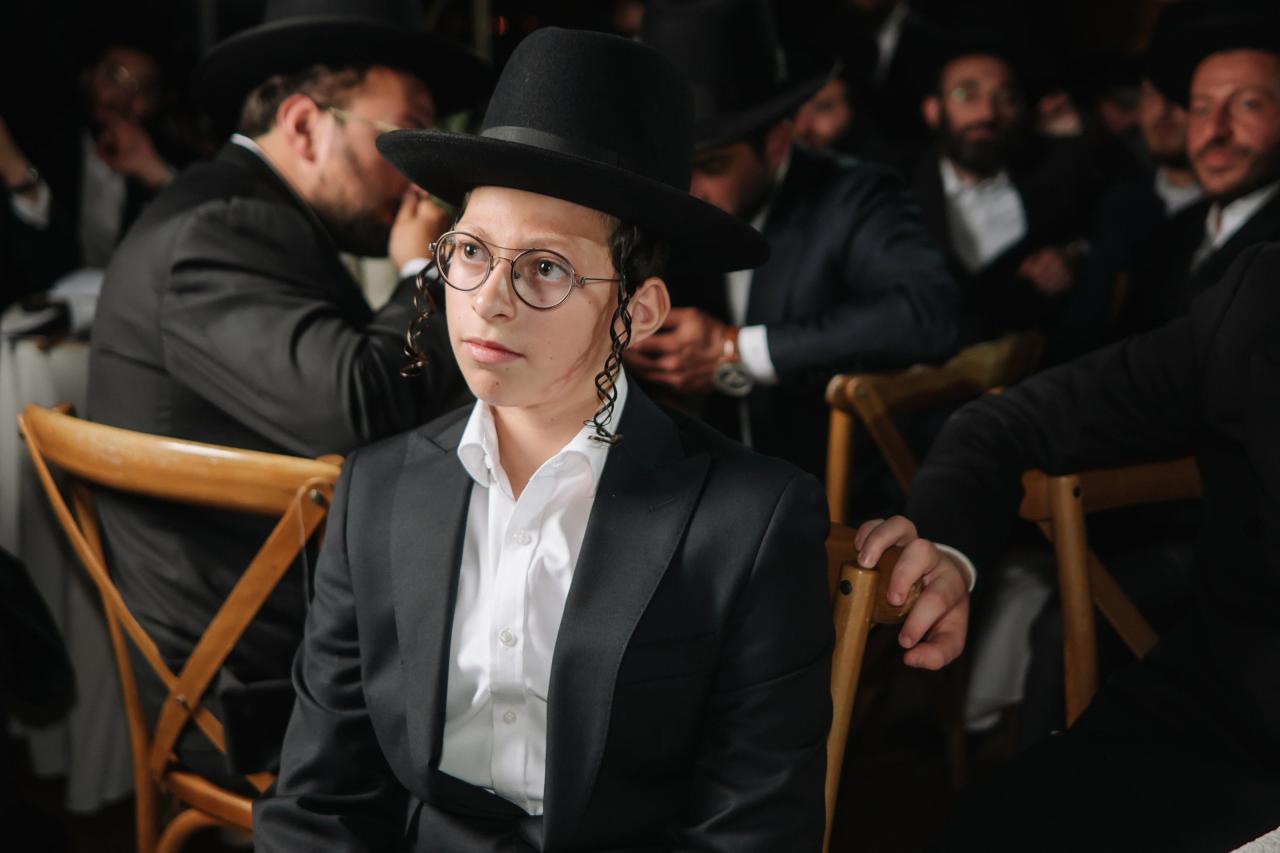 Bar Mitzvah boy in a black hat and glasses seated in a banquet hall during the celebration, with guests blurred in the background. Bar Mitzvah photography at The Arches, Brooklyn, NY.