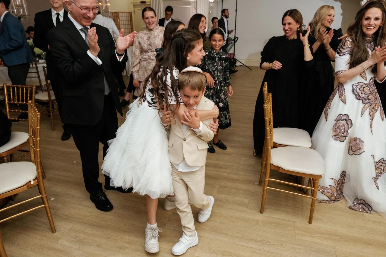 Candid dance-floor moment at a Jewish celebration: a little girl in a white outfit is held while family members clap and smile around her in a warmly lit hall. Wedding photography at Falkirk Estate Country Club, Central Valley, NY.
