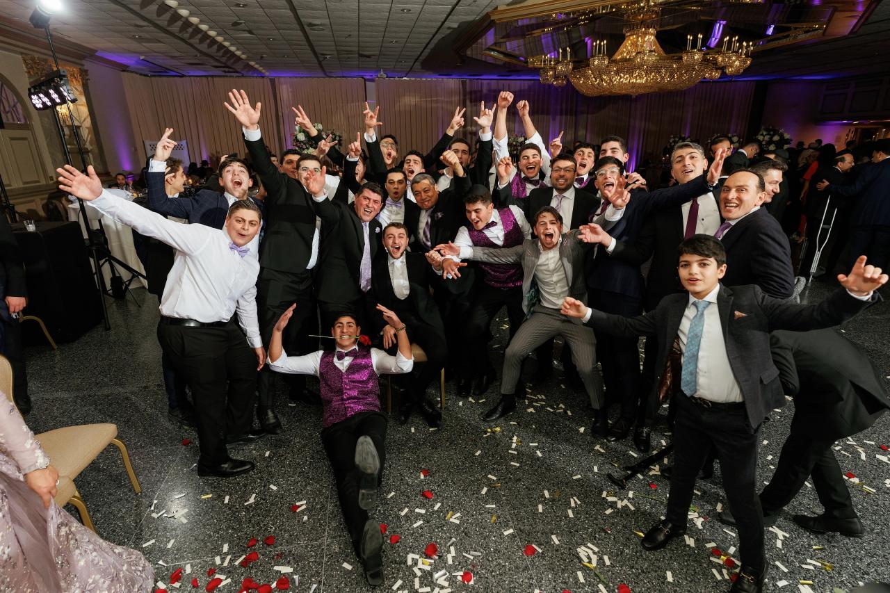 A group of boys and men at a wedding dance with arms raised and smiling broadly, celebrating with confetti on the floor in a ballroom.
