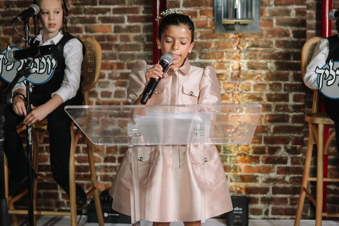 Young guest in a satin dress speaking into a microphone behind a clear podium against a brick wall, with other children seated or standing beside the stage area. Bar Mitzvah photography at The Arches, Brooklyn, NY.