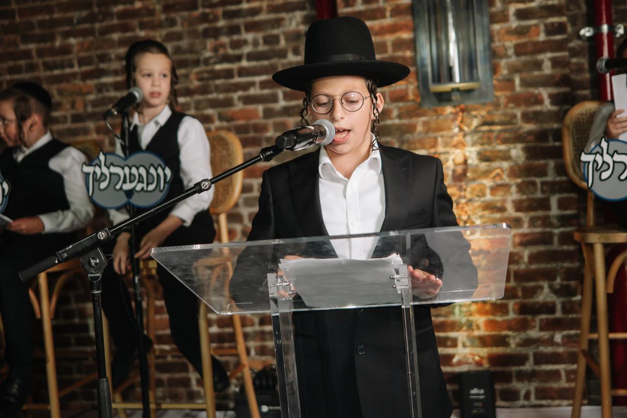 Young boy in formal attire wearing a black hat and glasses speaking into a microphone at a clear podium against a brick wall backdrop at a celebration. Bar Mitzvah photography at The Arches, Brooklyn, NY.