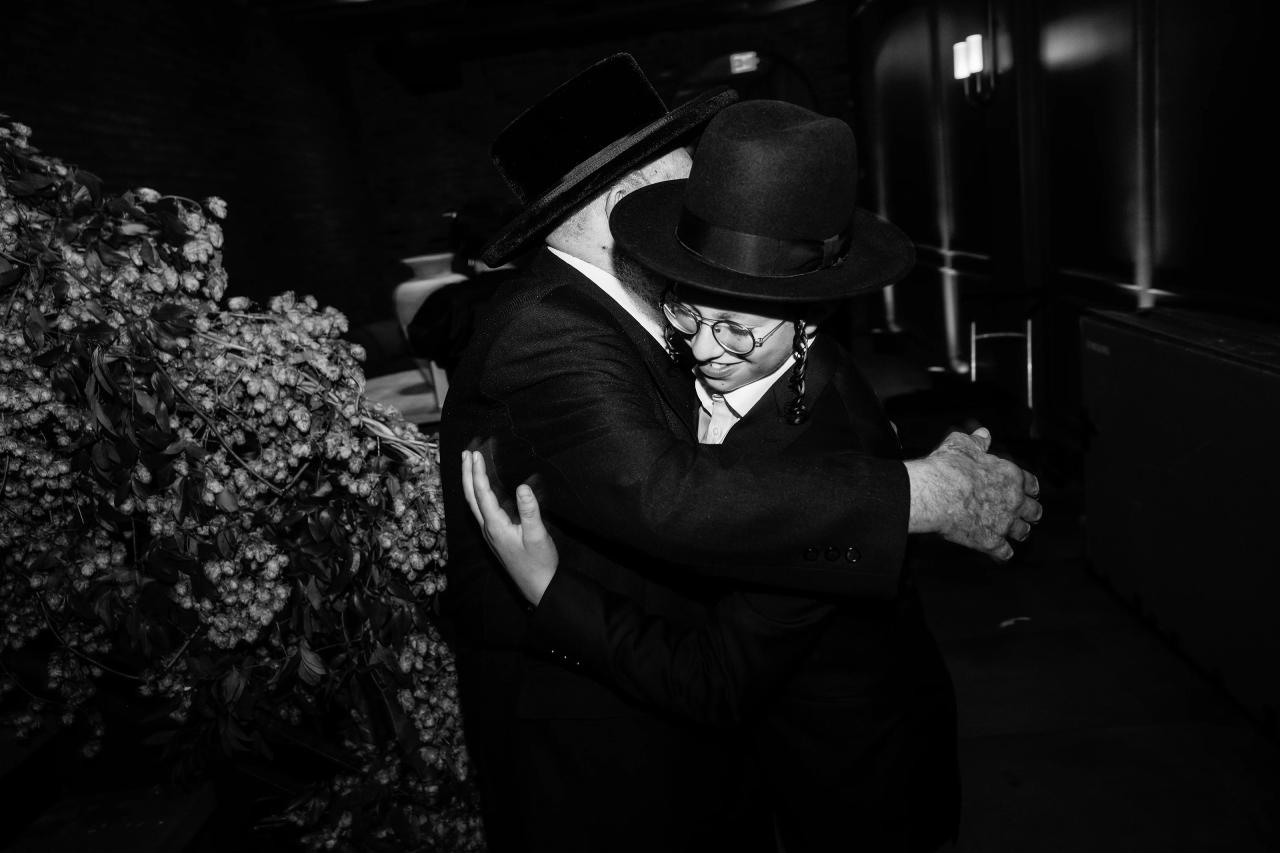 Black-and-white reception photo of a man in a black hat and suit hugging beside a large floral arrangement, smiling in a warm, intimate moment. Bar Mitzvah photography at The Arches, Brooklyn, NY.