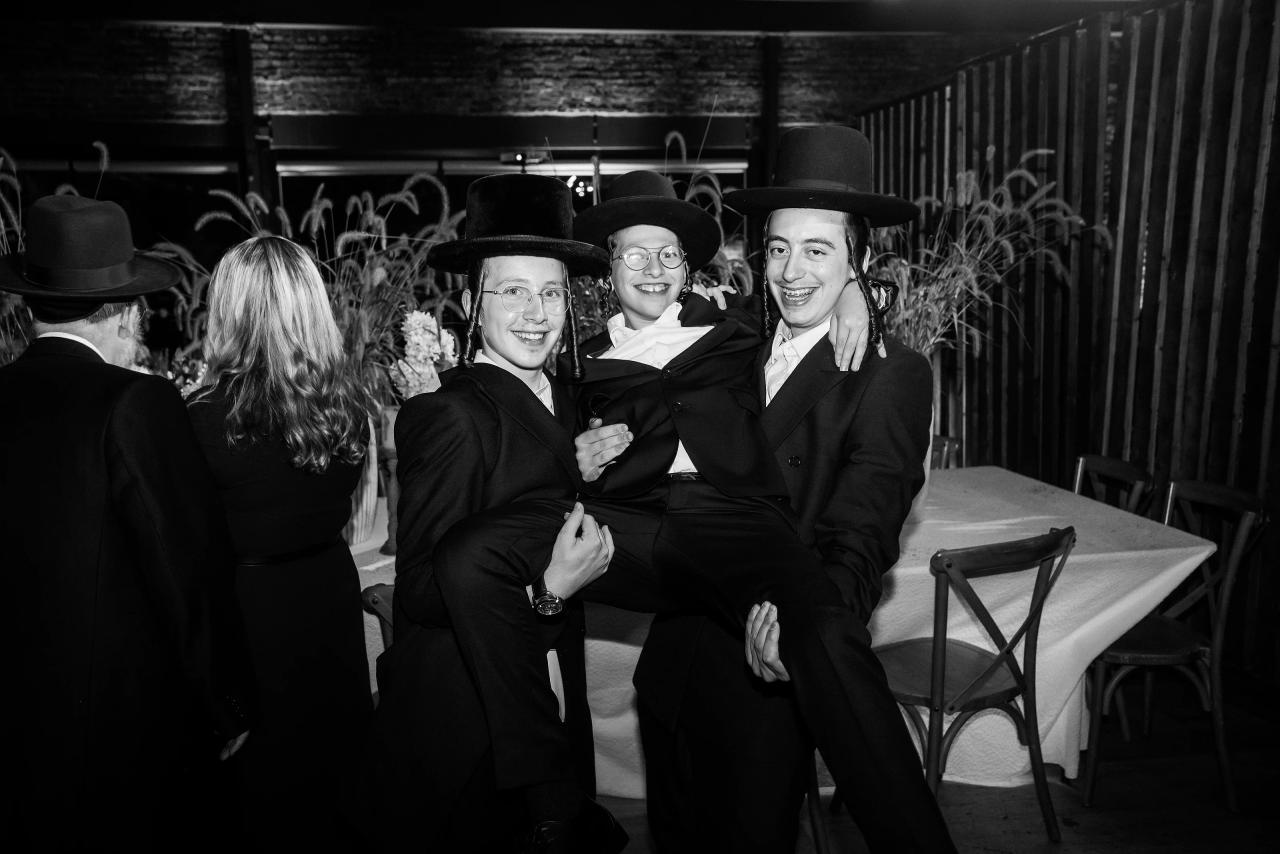 Black-and-white photo of three celebrants in formal black outfits and hats sharing laughter during a Bar Mitzvah celebration, with a woman in the foreground facing away. Bar Mitzvah photography at The Arches, Brooklyn, NY.
