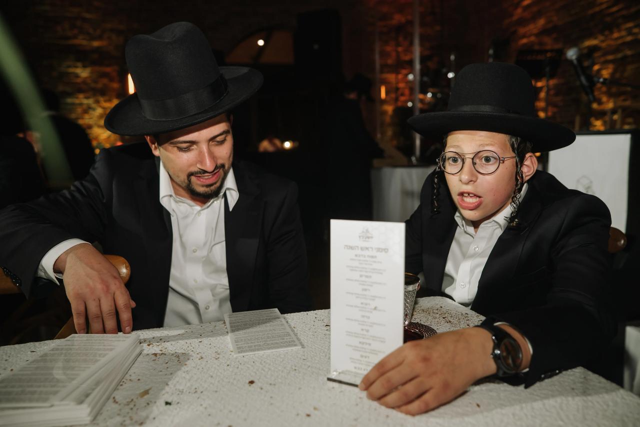 Two men in formal hats seated at a reception table, one reading papers while the other looks up with a surprised expression; warm indoor lighting and confetti on the table. Bar Mitzvah photography at The Arches, Brooklyn, NY.
