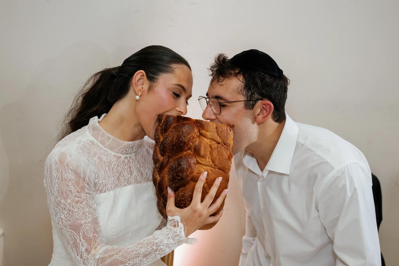 Frum wedding couple sharing an intimate kiss while holding bread, close-up against a plain light background. Wedding photography at Falkirk Estate Country Club, Central Valley, NY.