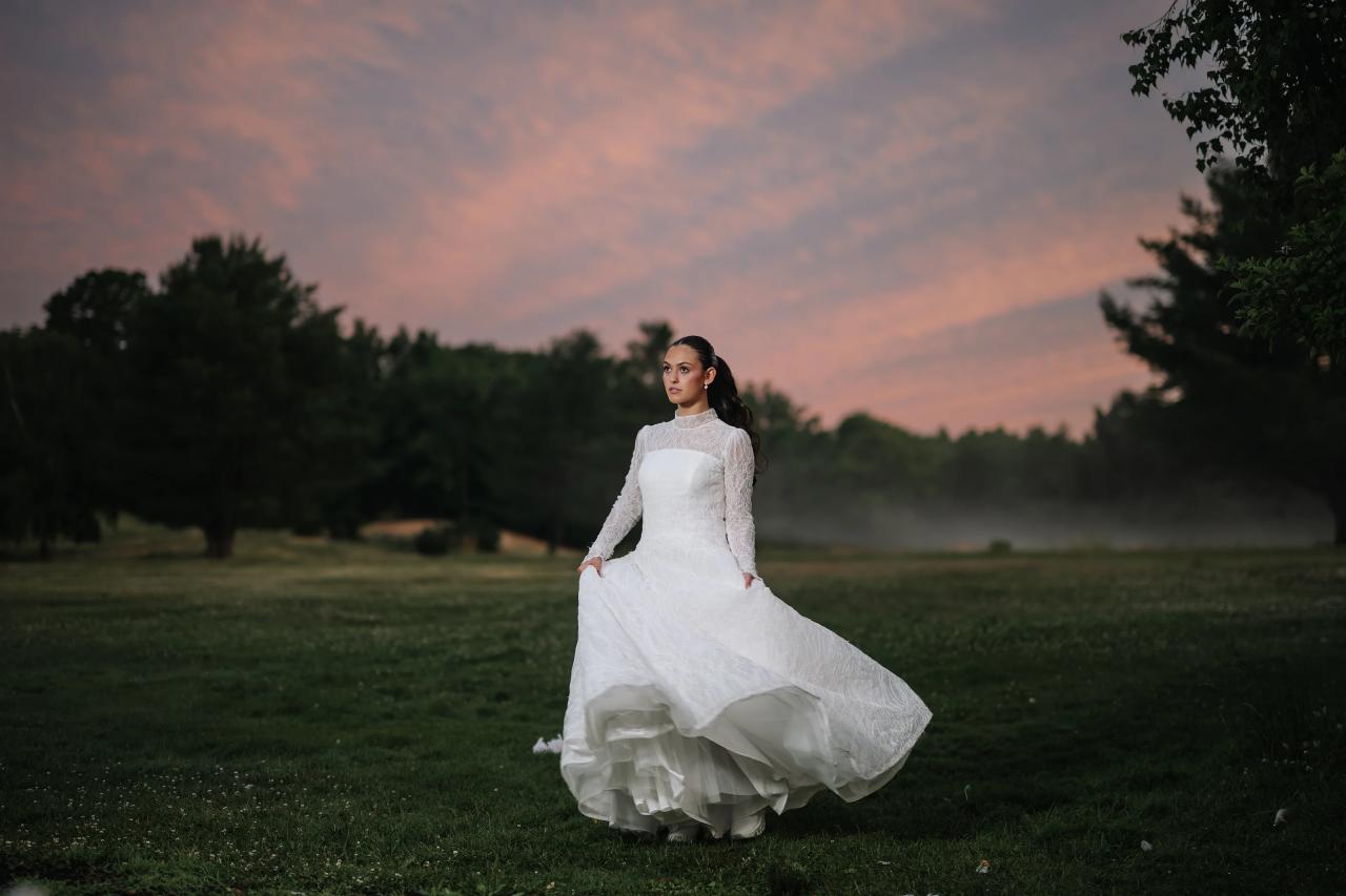 Bride in a white lace wedding gown walking across a grassy field at sunset, her skirt flowing, with a pink twilight sky and trees in the background. Wedding photography at Falkirk Estate Country Club, Central Valley, NY.