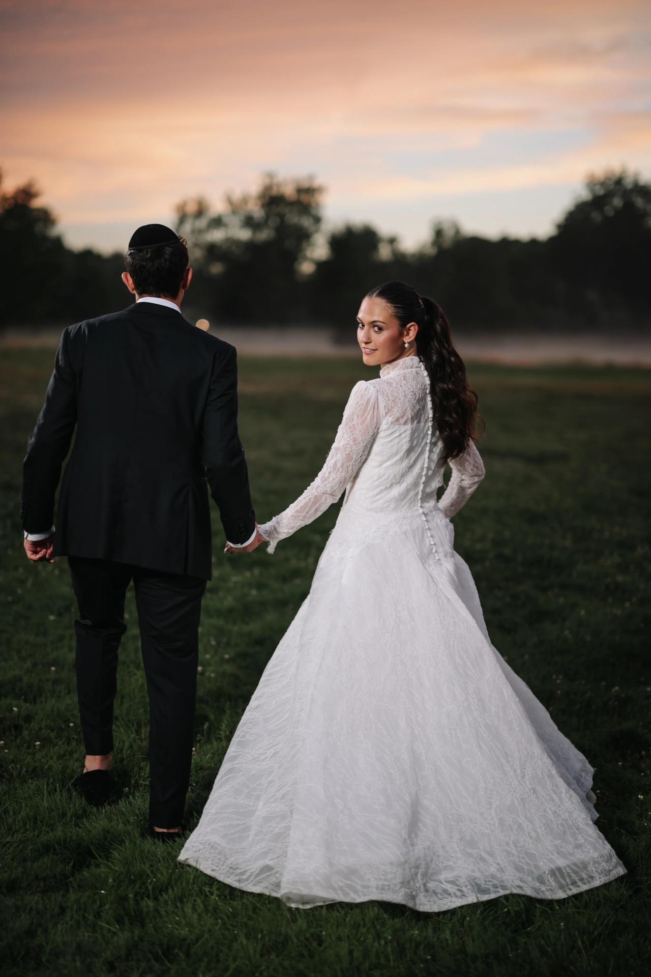 Bride in a white gown walking hand-in-hand with groom in a black suit on a grassy lawn at sunset, looking over her shoulder with warm sky behind them. Wedding photography at Falkirk Estate Country Club, Central Valley, NY.
