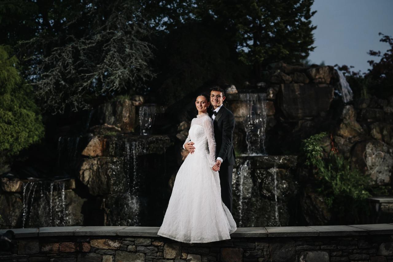 Romantic wedding portrait of a bride and groom standing together in front of a nighttime fountain and stone wall with greenery, holding hands. Wedding photography at Falkirk Estate Country Club, Central Valley, NY.
