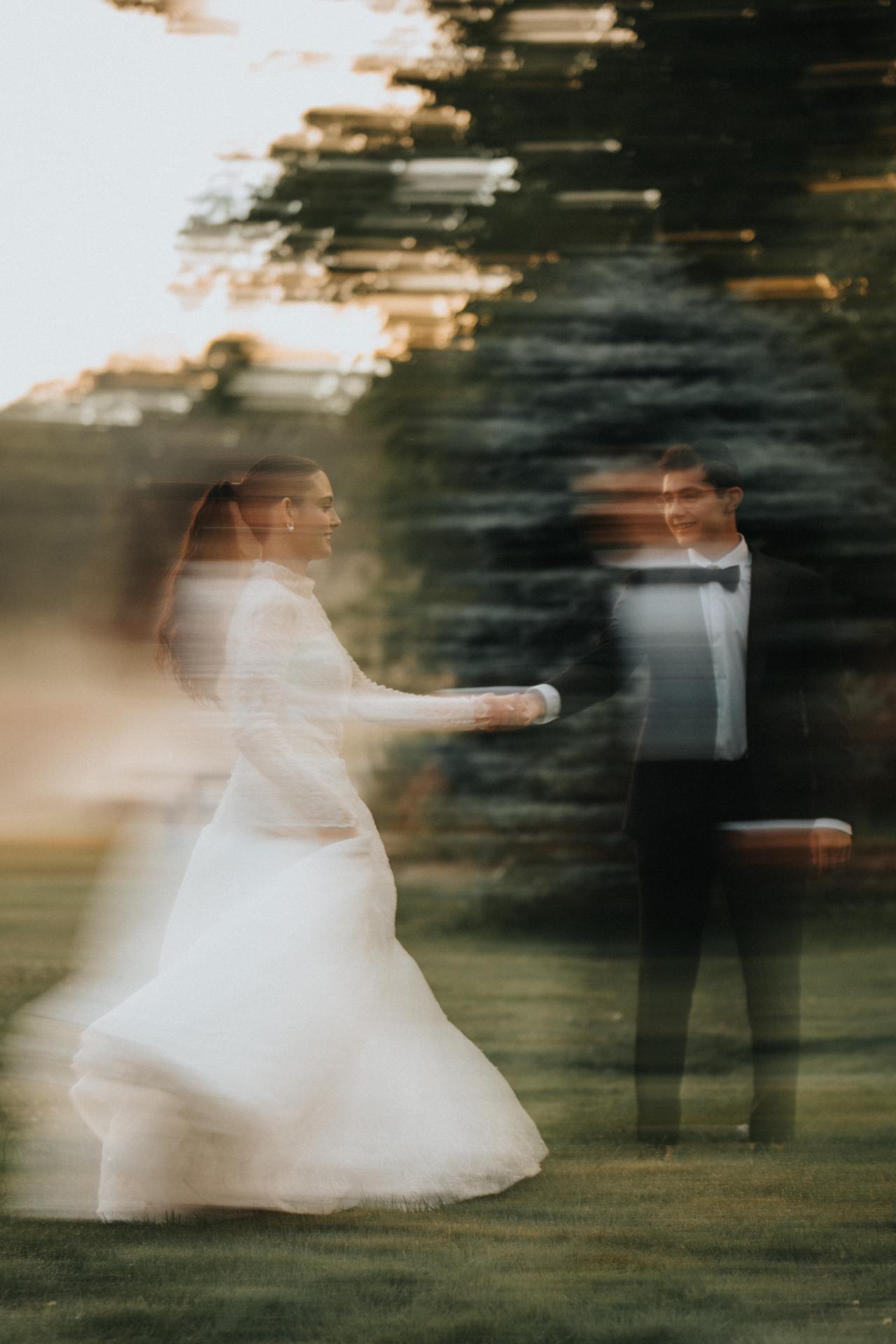 The bride in a white wedding dress and the groom in formal attire captured with long-exposure blur as they walk together outdoors at sunset, with greenery behind them. Wedding photography at Falkirk Estate Country Club, Central Valley, NY.