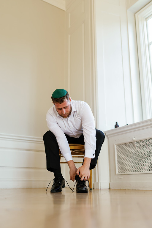 A man wearing a green kippah and white shirt kneels indoors while adjusting and tying his shoes before the event.
