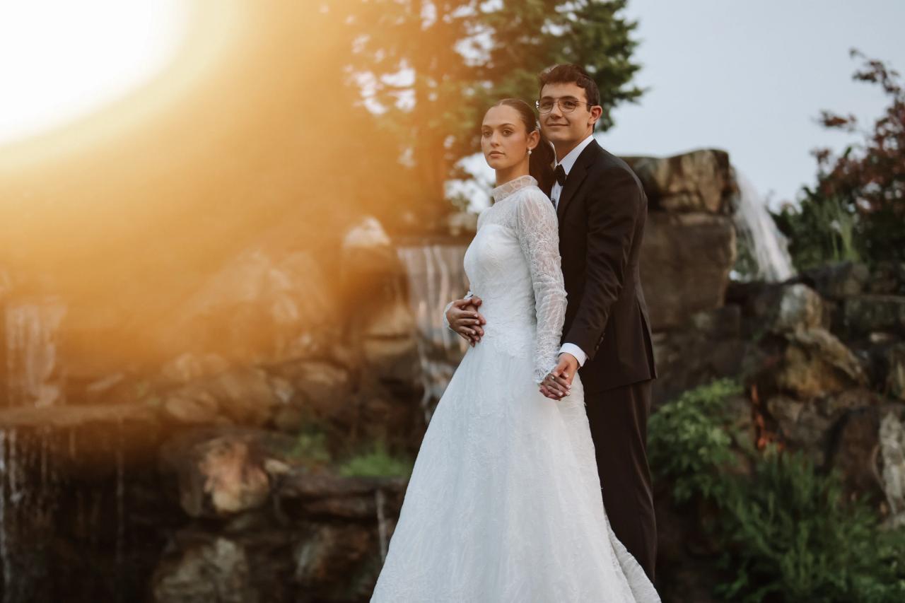 Golden-hour outdoor wedding portrait of a bride and groom walking together, framed by greenery and a warm sun flare from the left. Wedding photography at Falkirk Estate Country Club, Central Valley, NY.
