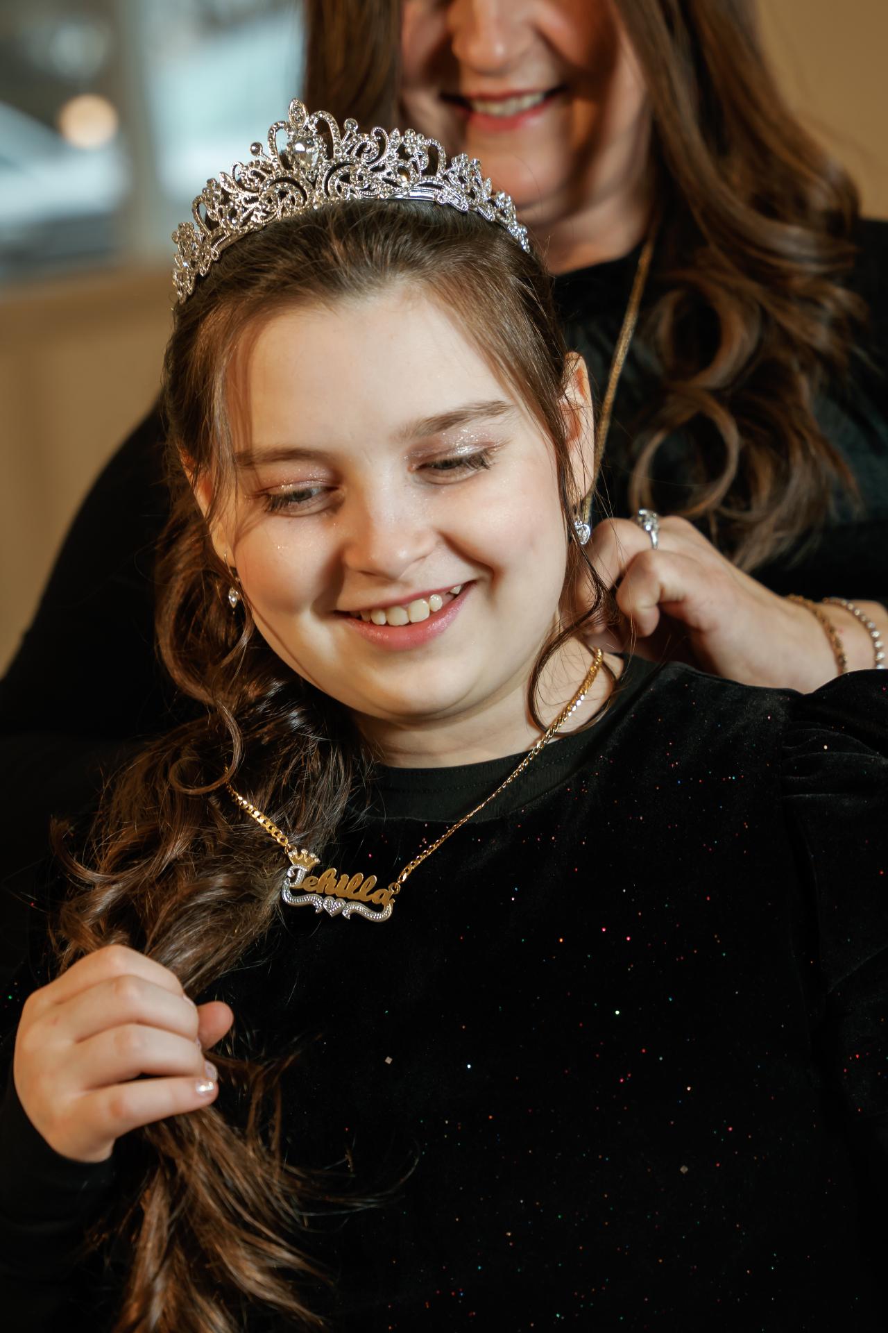 Smiling girl wearing a tiara and a necklace, with a softly blurred warm background at an event.