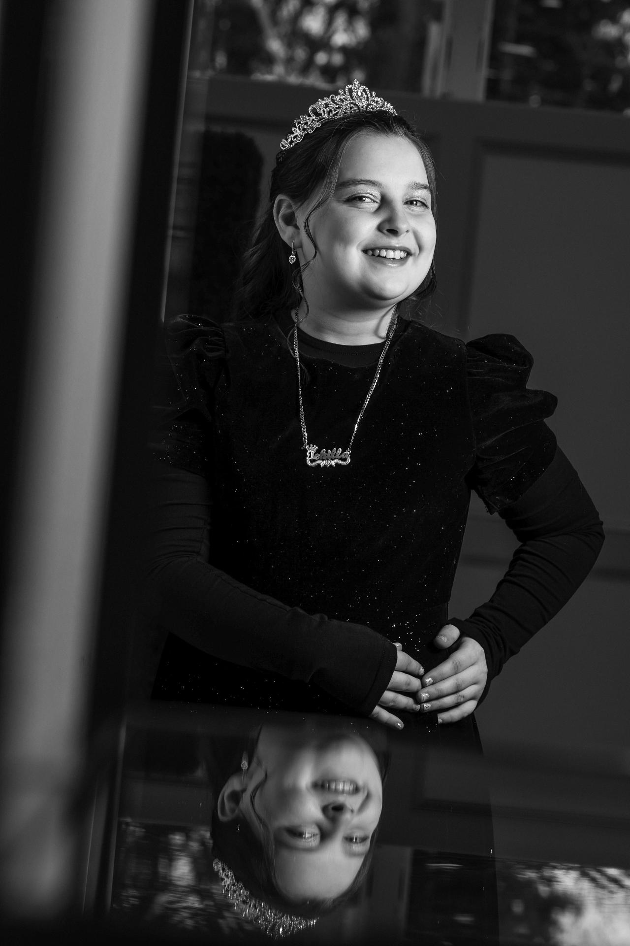 Black-and-white portrait of a joyful girl wearing a tiara and formal dark outfit, with a subtle reflection beneath her in the frame.