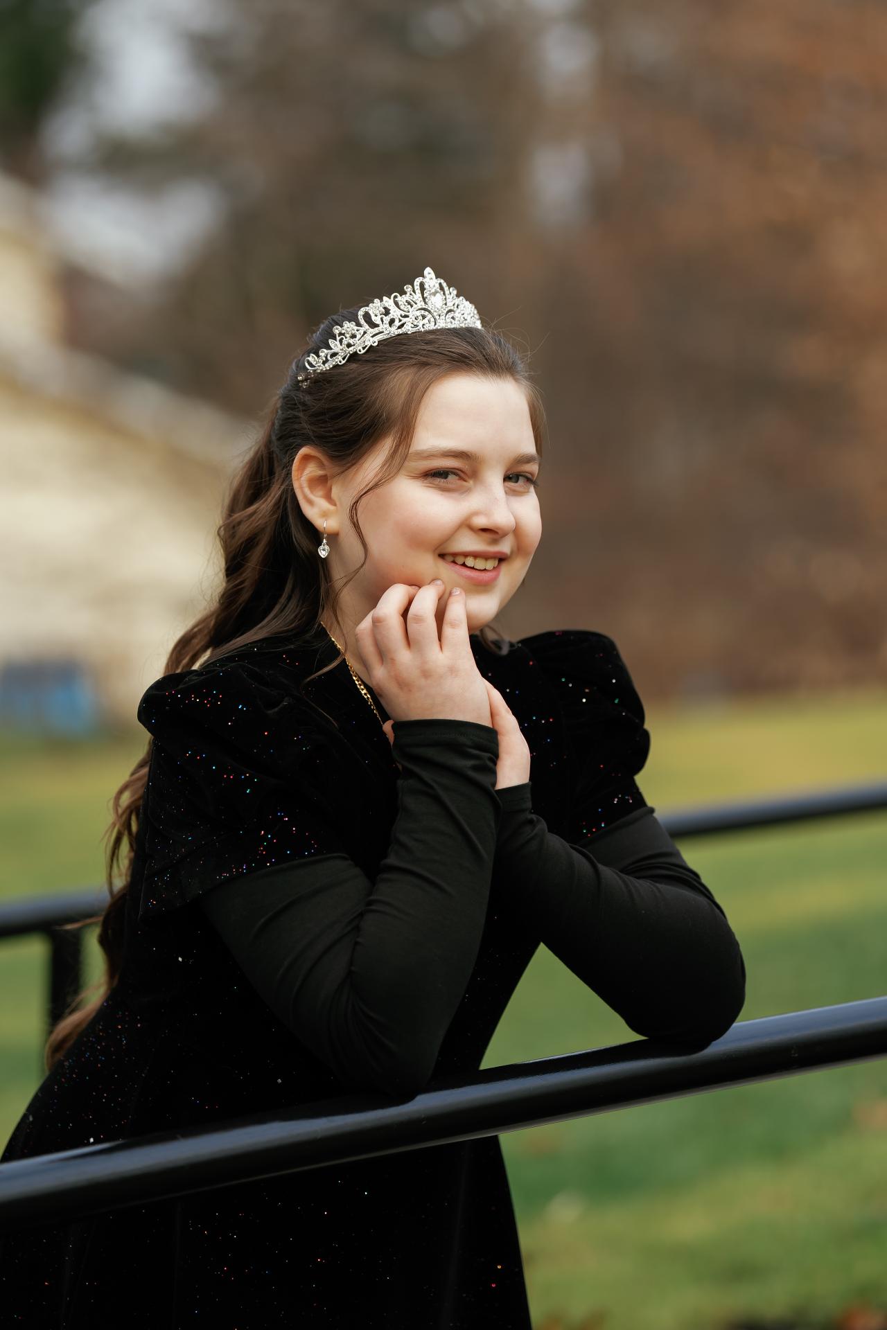 Smiling young girl in a tiara and black outfit posing outdoors with her hands near her cheek, against a soft green background.